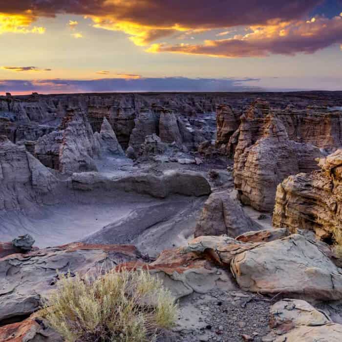 Rock formations and canyons under a colorful sunset sky with sparse vegetation in the foreground.