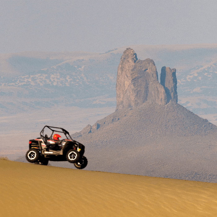 A person drives an off-road vehicle across sand dunes with a rocky hill and hazy mountains in the background.