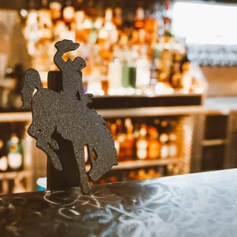 A metal silhouette of a cowboy riding a bucking horse stands on a bar counter, with shelves of liquor bottles blurred in the background.