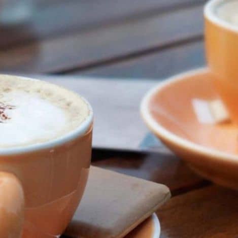 Two beige coffee cups on saucers sit on a wooden table, one filled with a foamy drink and the other partially out of frame.