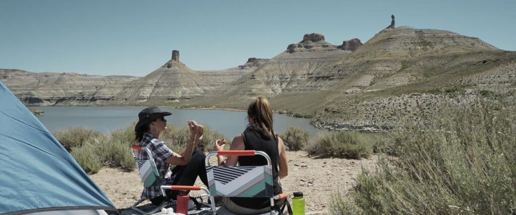 Two people sit in folding chairs by a tent near a lake, with rocky hills and sparse vegetation in the background under a clear sky.