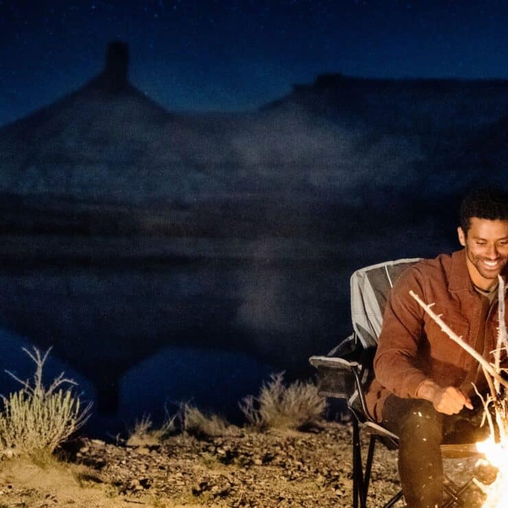 Two people sit by a campfire near a lake at night, with mountains silhouetted in the background under a starry sky.