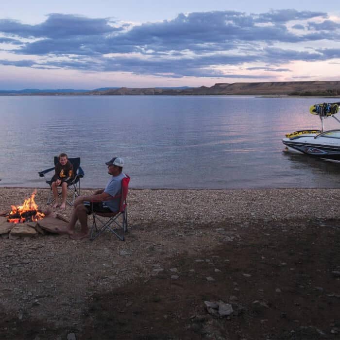 Two people sit in camping chairs by a campfire on a rocky lakeshore at dusk, with a boat docked nearby and calm water in the background.