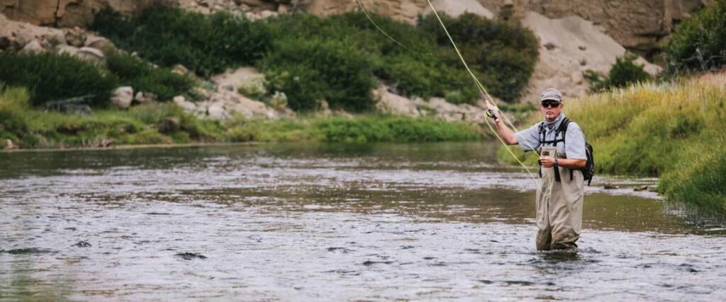 Person wearing waders stands in a shallow river fly fishing with a rod, surrounded by greenery and rocky terrain.