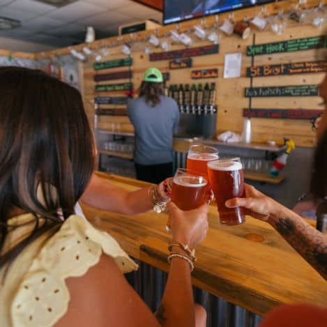 Three people clink glasses of beer at a wooden bar, with a bartender in the background and beer taps and menu boards visible on the wall.