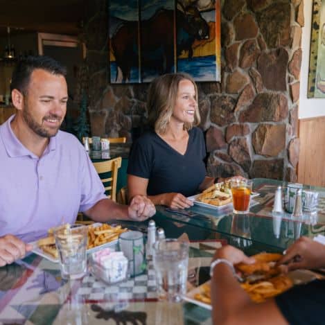 Two people sit at a restaurant table with sandwiches, fries, and drinks, smiling and talking to a third person whose arm is visible in the foreground.