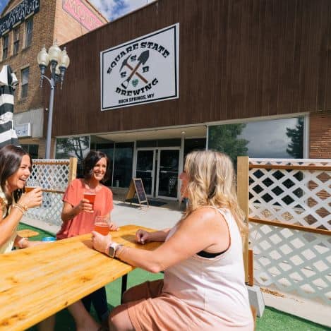 Three women sit at a wooden table outdoors, drinking and talking in front of Square State Brewing in Rock Springs, WY.
