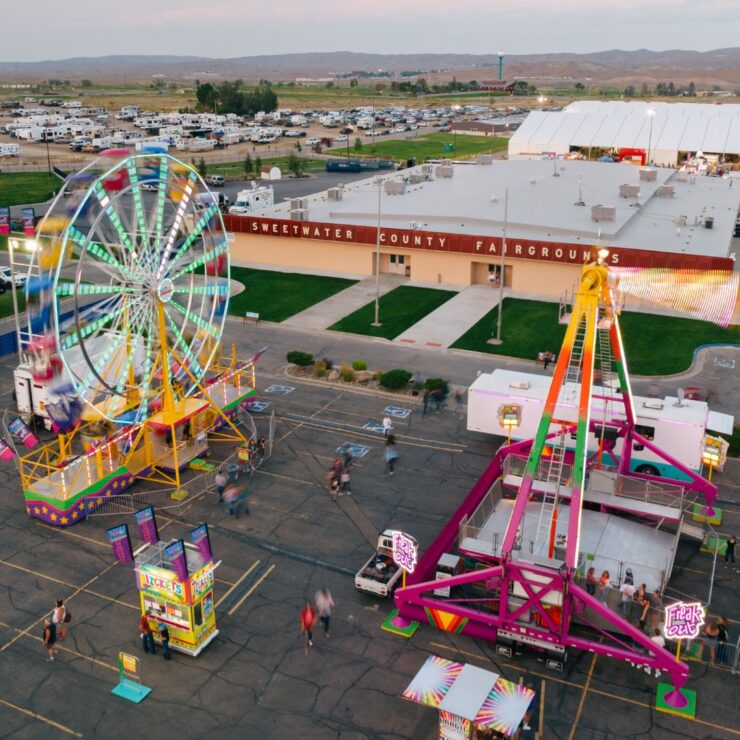 Aerial view of a county fairground with a Ferris wheel, amusement rides, scattered visitors, parked cars, and the Sweetwater County Fairgrounds building in the background.