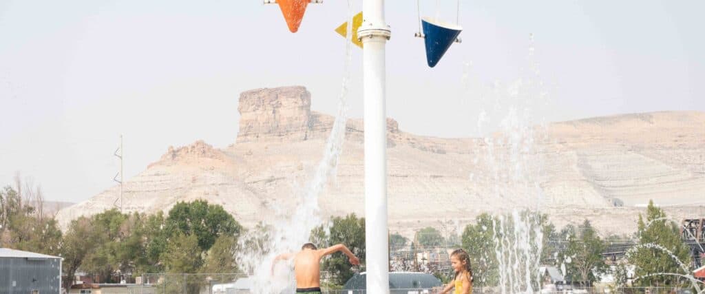 Two children play under water buckets at a splash pad, with desert cliffs and trees visible in the background.