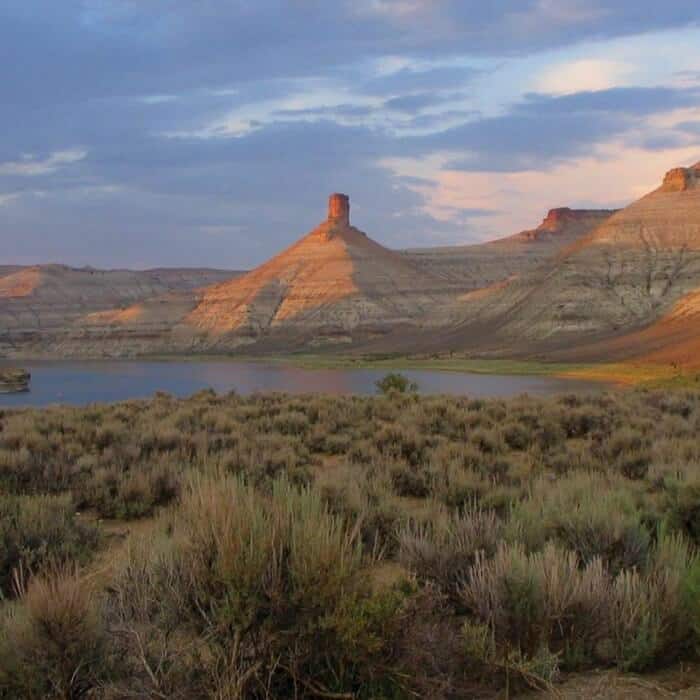 Desert landscape with dry shrubs in the foreground, a calm lake, and rocky mesas under a partly cloudy sky at sunset.