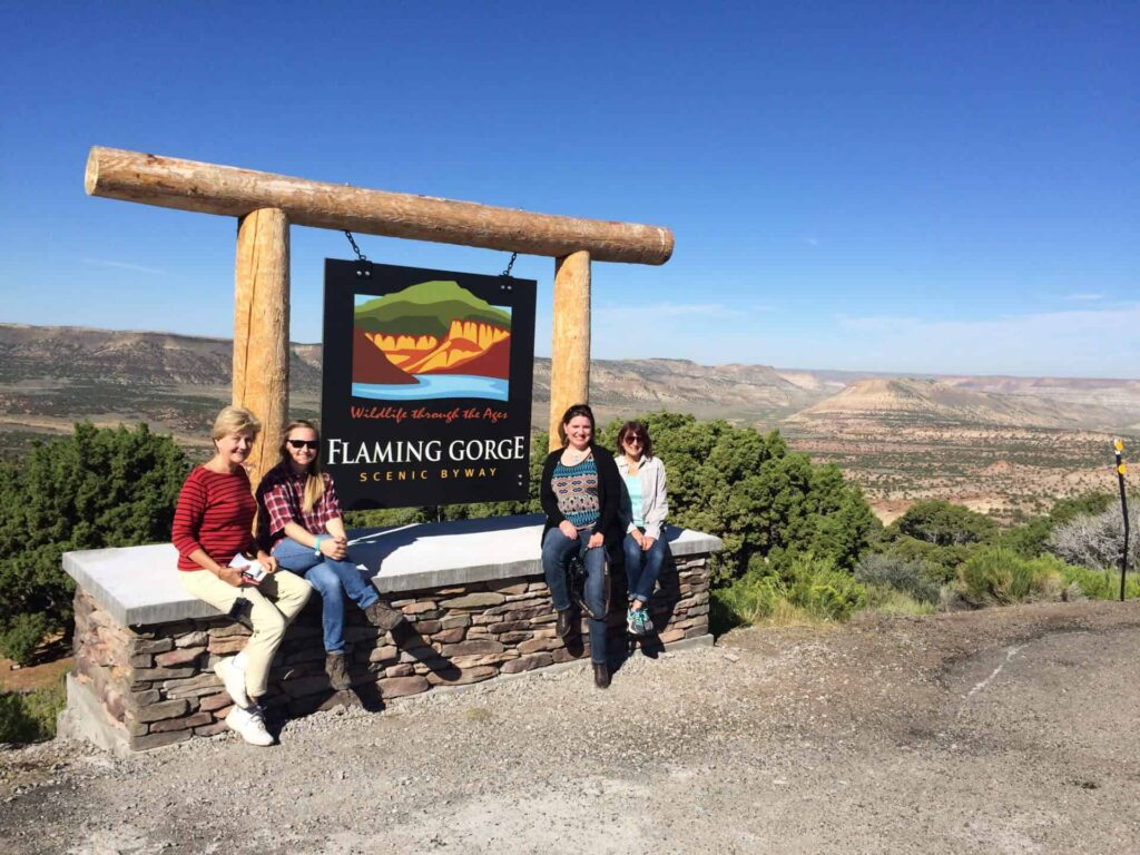 Four people sit on a stone ledge beneath a large wooden sign reading "Flaming Gorge Scenic Byway," with a scenic desert landscape in the background.