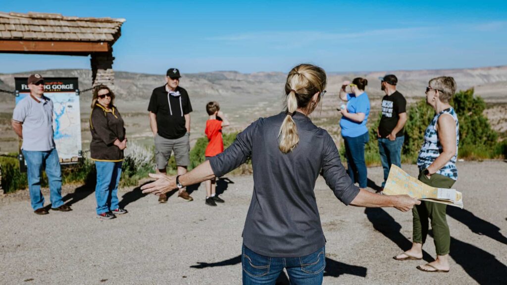 A woman holding a map addresses a small group of people outdoors near an informational sign, with mountains visible in the background.