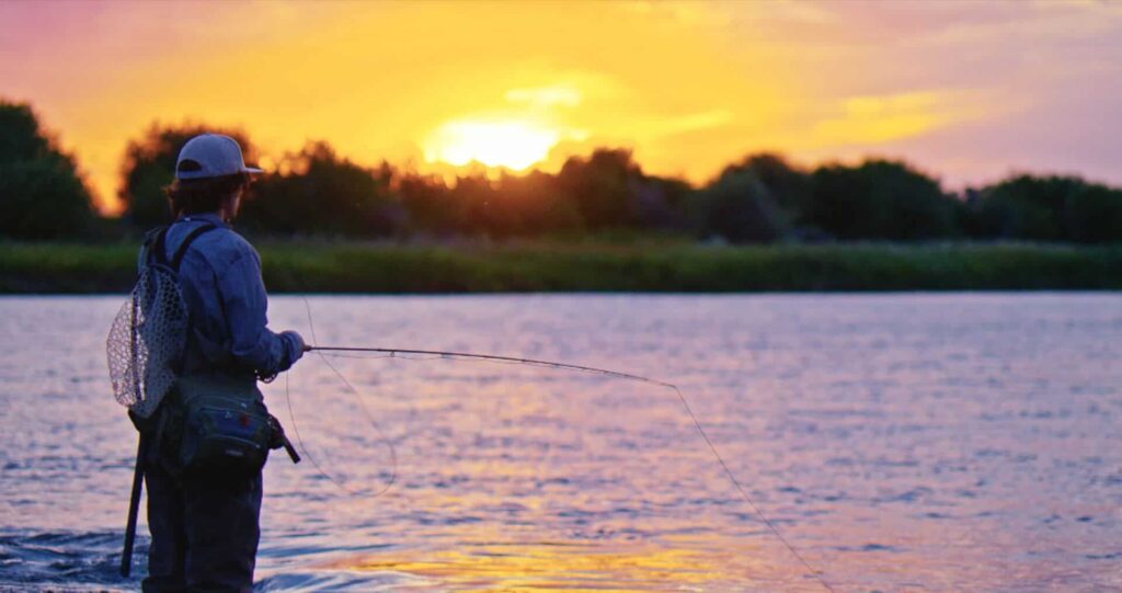 A person stands in shallow water fishing with a rod at sunset, wearing outdoor gear and a cap, with trees visible in the background.