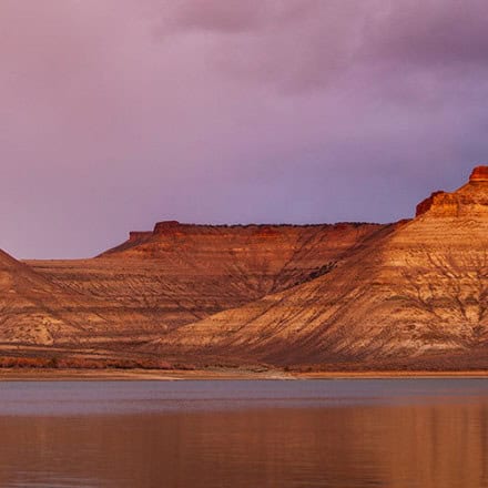 A calm body of water reflects layered, reddish-brown mesas and hills under a purple and pink sky.