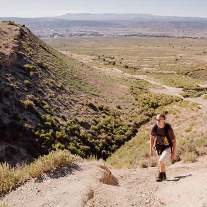 A person wearing a backpack hikes up a dusty trail in a dry, hilly landscape with distant mountains and a town visible in the background.