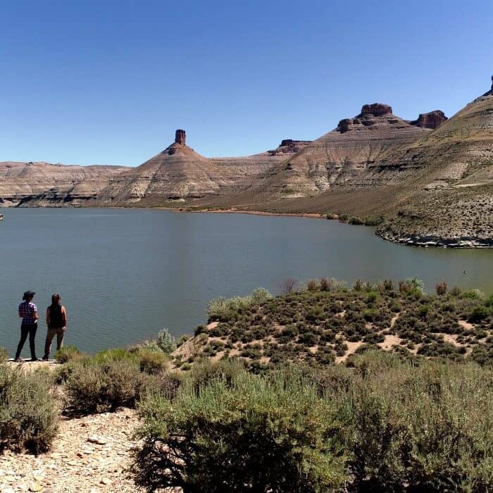 Two people stand on a rocky shore overlooking a wide river or lake, surrounded by arid hills and mesas under a clear blue sky.