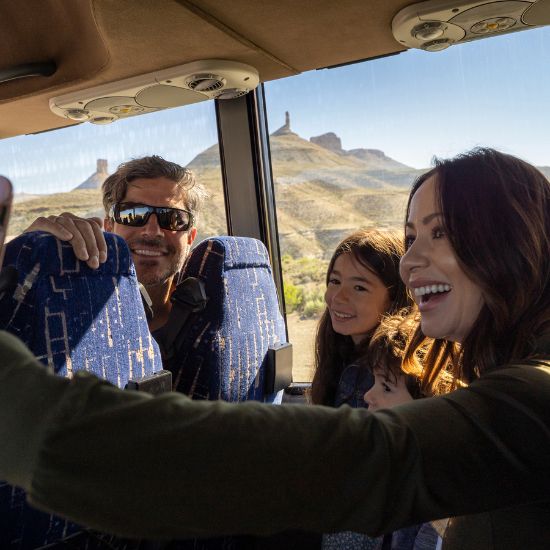A group of people sit together on a bus, smiling and taking a selfie, with mountains visible through the window in the background.