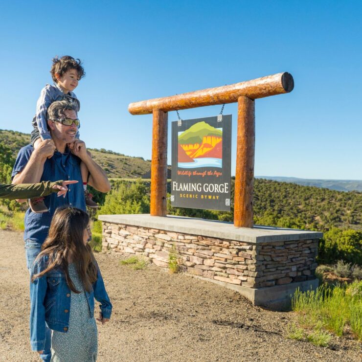 A family walks near a sign for Flaming Gorge Scenic Byway surrounded by green hills and a clear blue sky.