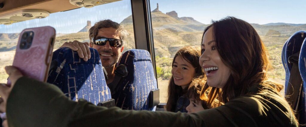 A group of people sit on a tour bus, smiling and taking a selfie with desert hills and rock formations visible through the window.