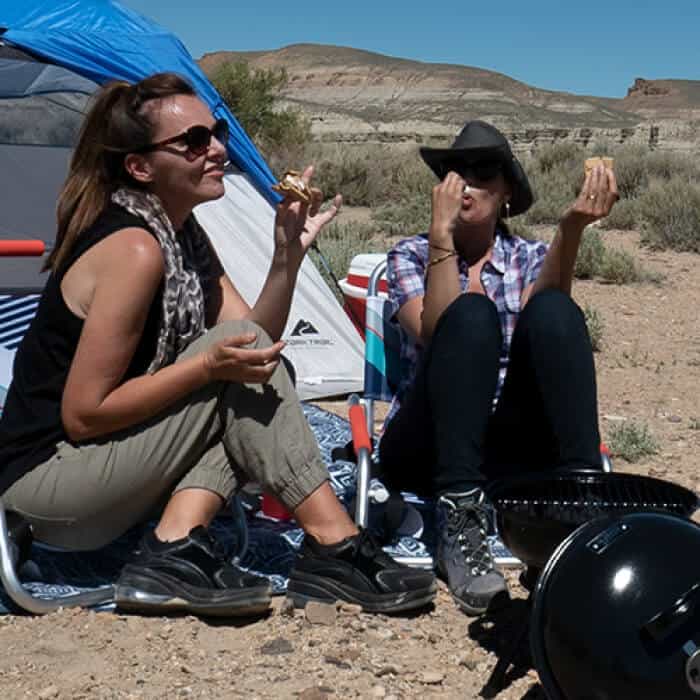 Two women sit on camping chairs by a tent in a desert area, eating food and talking, with a portable grill nearby under a clear sky.
