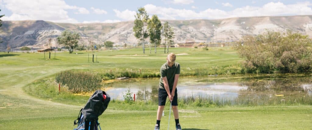 A person stands in a golf stance with a club on a green golf course near a pond, with hills and trees in the background. A golf bag sits nearby.