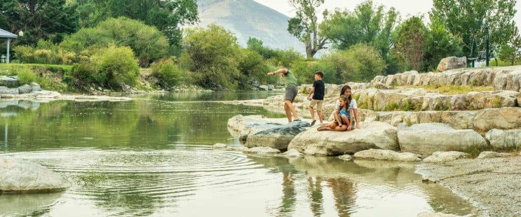 Four children are playing by a river, with two standing on rocks near the water and two sitting on large stones. Trees and hills are in the background.