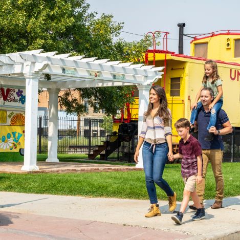 A family of four walks outside near a yellow train car and a white pergola on a sunny day.