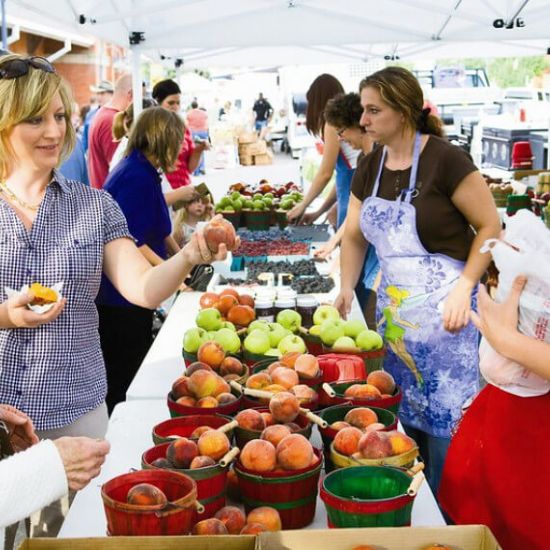 People shop for peaches, apples, and berries at an outdoor farmers market. Vendors stand behind tables displaying baskets of fruit under white tents.