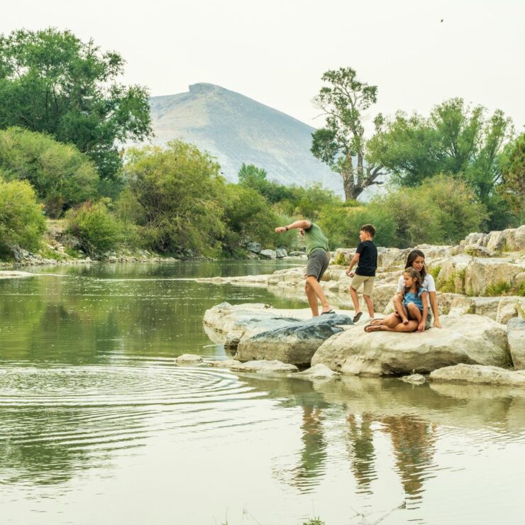 Three children play on rocks beside a calm river, with trees and a mountain in the background on a clear day.
