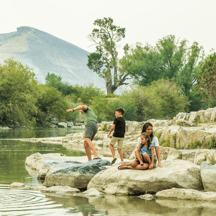 Four people are on rocks by a river; one person skips stones while the others sit or stand nearby, with trees and a mountain in the background.