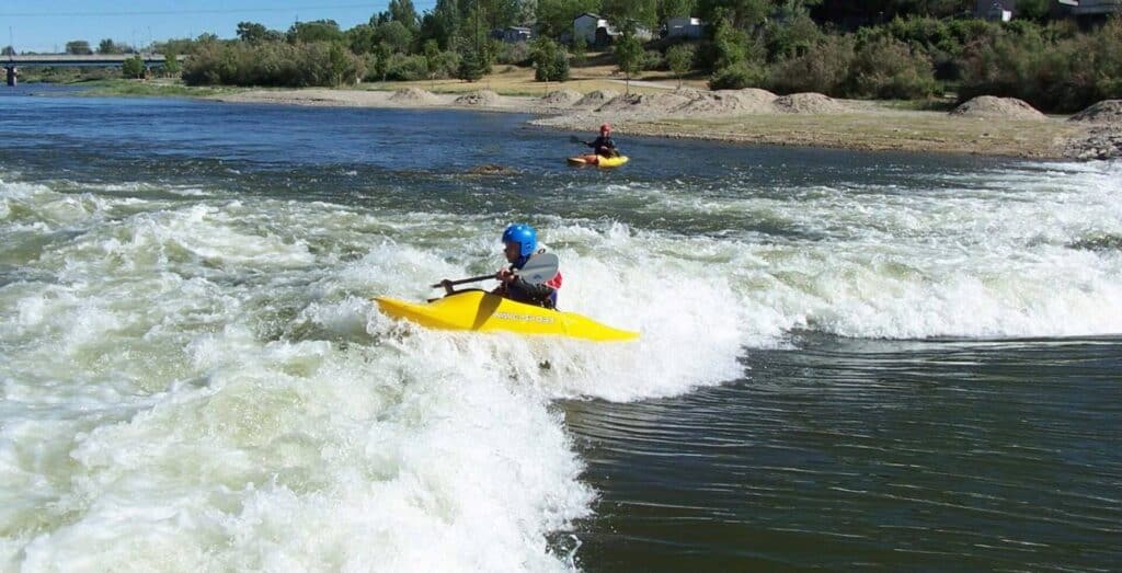 Two people in yellow kayaks paddle through river rapids, with one closer to the foreground and both wearing helmets and life jackets. Trees and sand are visible along the riverbank.