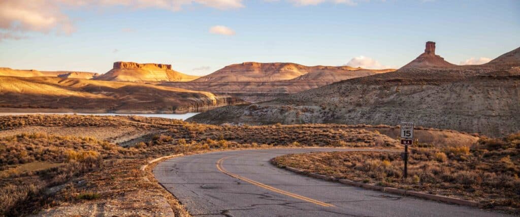 A winding road curves through a dry, rugged desert landscape with mesas and rock formations under a partly cloudy sky; a speed limit sign reads 15 mph.