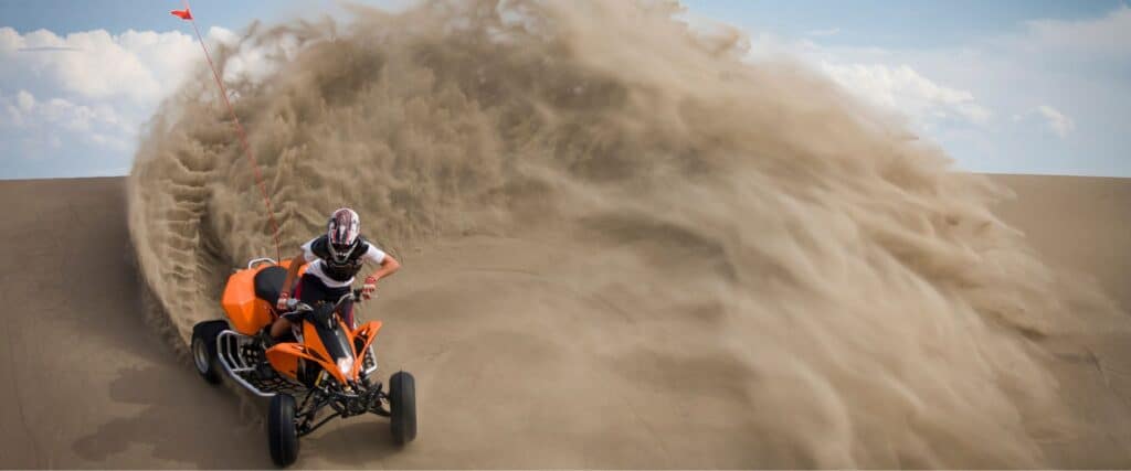 Person riding an orange ATV on sand dunes creates a large spray of sand behind them under a partly cloudy sky.