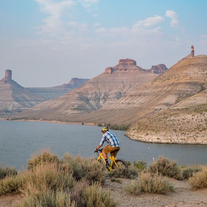 A person wearing a helmet rides a yellow bicycle on a trail overlooking a river and rocky desert mountains under a partly cloudy sky.