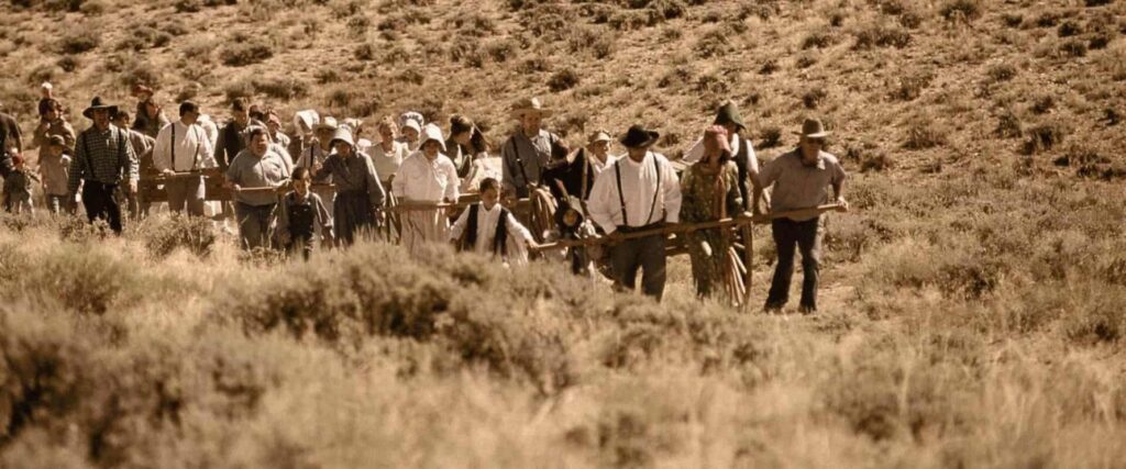 A group of people in pioneer-era clothing walk through dry, grassy terrain, pulling and pushing wooden handcarts.