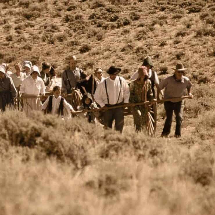 A group of people in pioneer-era clothing walk through a dry, grassy landscape, pulling handcarts and carrying supplies.