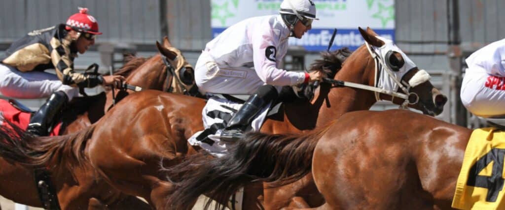 Jockeys riding racehorses in a close competition on a racetrack, with horses and riders in mid-stride.