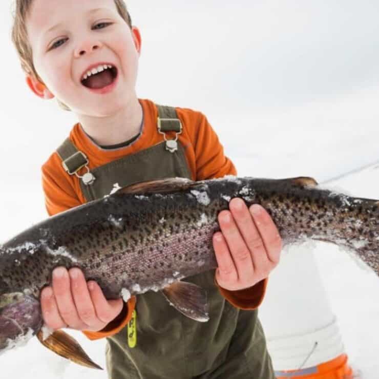 A smiling child in overalls holds a large fish with ice on its body, standing outdoors on a snowy surface.
