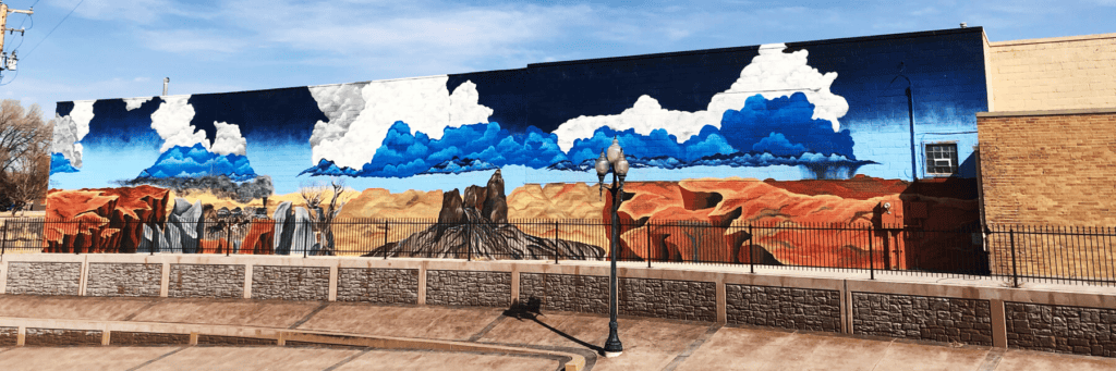 A mural on a brick wall depicts a desert landscape with red rocks, mesas, sparse vegetation, and blue and white clouds against a bright sky.