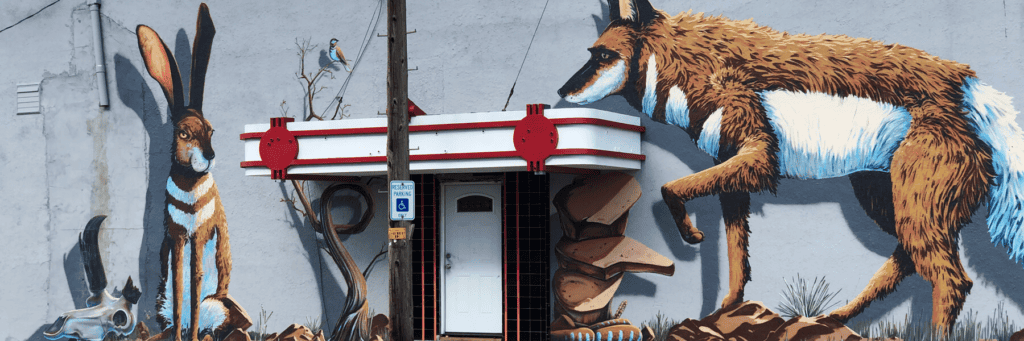 A mural shows a jackrabbit, a coyote, and desert scenery painted around a building entrance with a pole and accessibility sign in front.