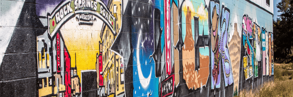 Colorful graffiti mural on a brick wall, featuring abstract designs and a section depicting a cityscape with a sign reading "Hot Springs" above it.