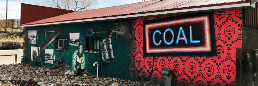 A mural on a building wall features mining tools, black and white photos, and a large neon-style “COAL” sign, with red and green patterned background. Rocks line the ground in front.