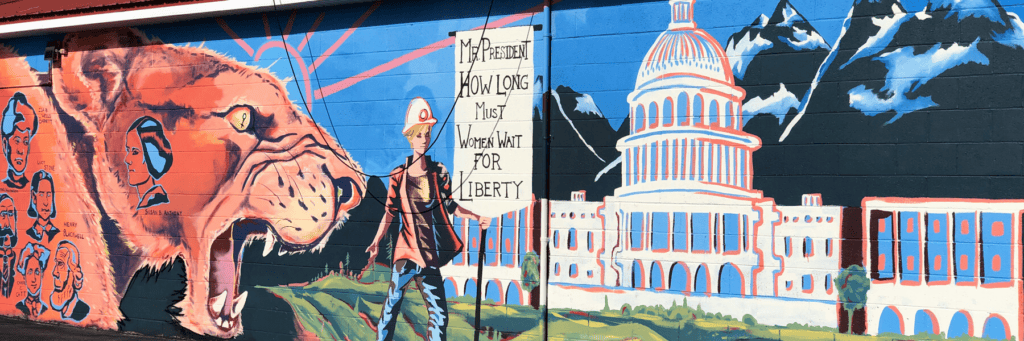 Mural of a roaring bear, a person holding a “How long must women wait for liberty” sign, faces, mountains, and the U.S. Capitol on an exterior wall.