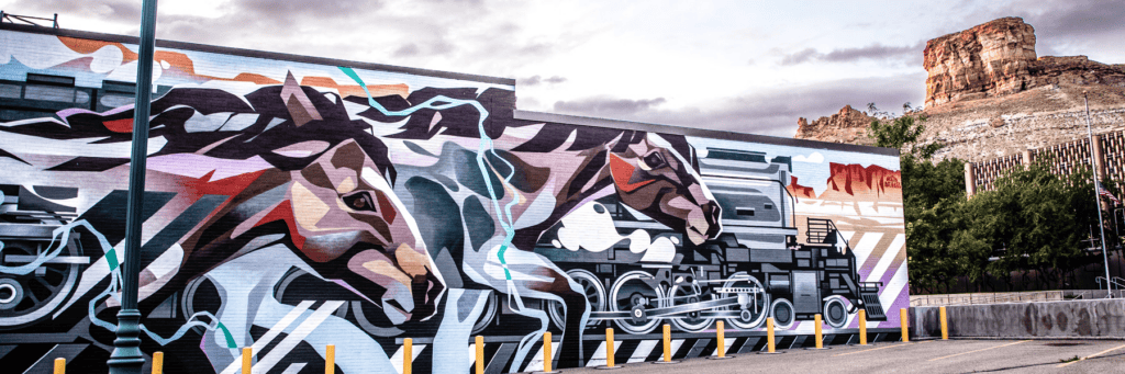 A mural of stylized running horses and mechanical elements covers a building wall, with a rocky hill and cloudy sky in the background. Yellow posts line the edge of the parking area.