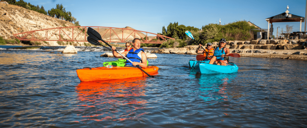 Three people in life jackets kayak on a river near a red bridge and stone shore, with trees and a gazebo in the background under a clear blue sky.