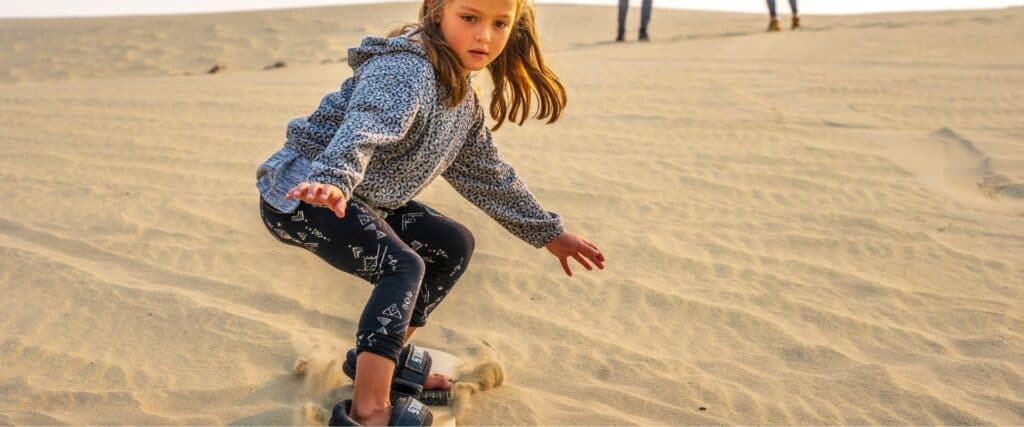 A young girl in patterned clothes sandboarding down a sandy dune, with two people standing in the background.