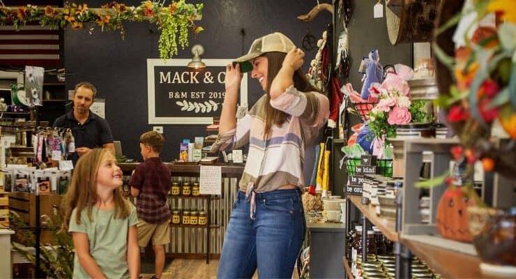 A woman tries on a hat in a store while a young girl smiles at her. A man and boy stand by the counter in the background. Shelves display various goods and decorations.