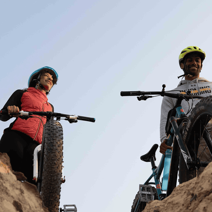 Two cyclists wearing helmets stand on rocks with their bikes, viewed from below against a clear sky.