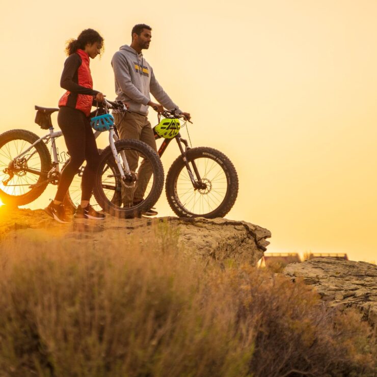 Two people stand with their fat-tire bikes on a rocky cliff at sunset, looking out over the landscape.