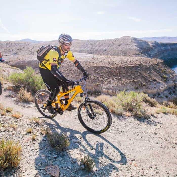 A cyclist wearing a yellow outfit and helmet rides a mountain bike on a rocky trail with hills and a canyon in the background.